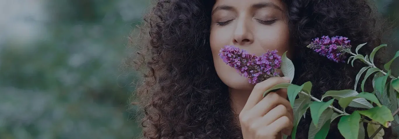 Frau mit lockigem dunklem Haar hält lila Blüten an ihr Gesicht, grünes Laub im Hintergrund.