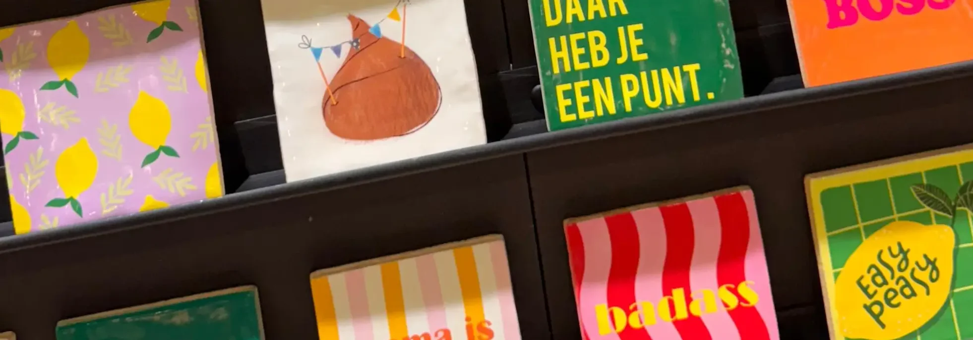 Bunte Postkarten und Kunstdrucke in schwarzem Regal: Zitronenmuster auf Violett, orangefarbener Ballon mit Wimpeln…