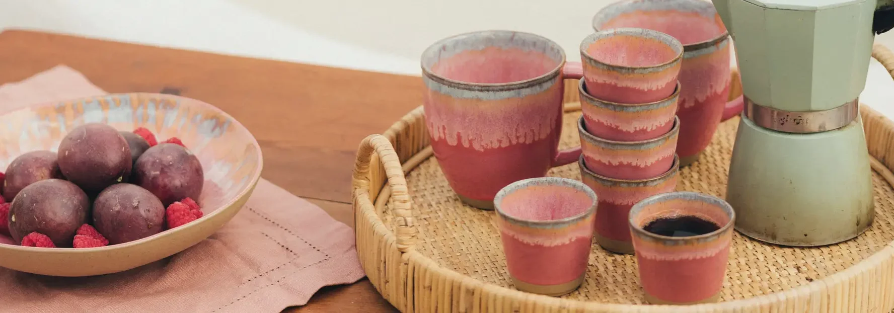 Rosa glasierte Keramiktassen und Becher auf einem geflochtenen Tablett, daneben eine Schale mit Früchten und Himbeeren auf…