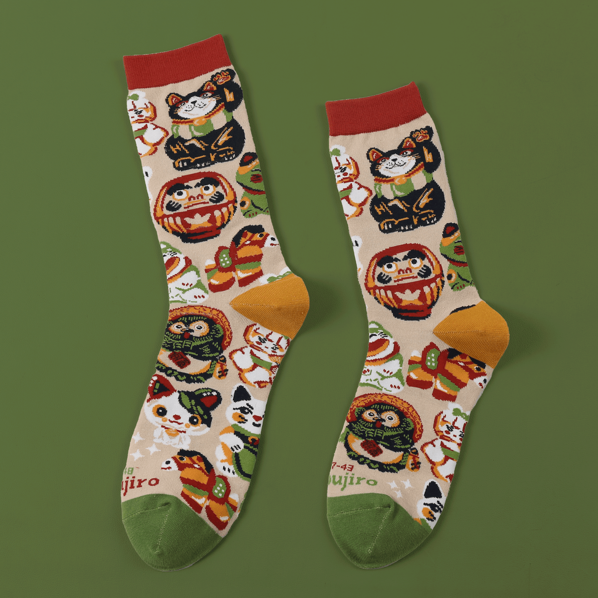 Zwei Socken nebeneinander auf grünem Grund: cremefarbenes Baumwollgewebe mit bunten japanischen Glücksbringer-Puppen in…
