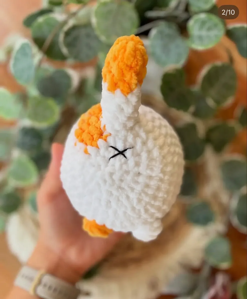 Gehäkeltes Amigurumi-Katzenplüschi in Weiß mit orangefarbenen Ohren und Pfoten, in einer Hand gehalten vor grünen…