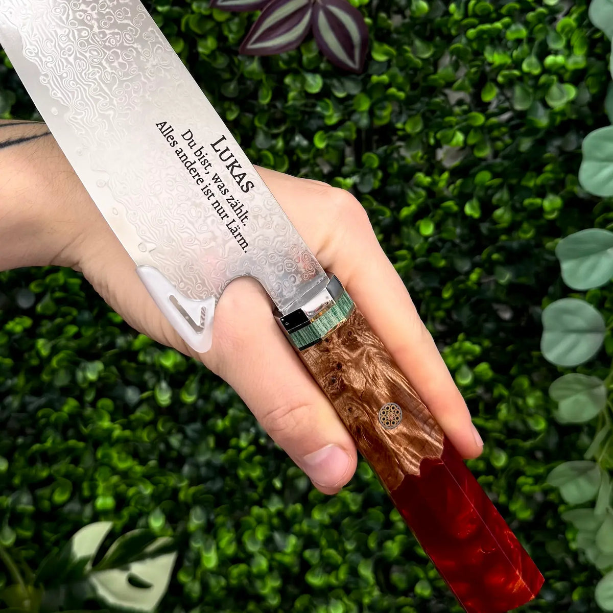 Damastmesser in Hand, Klinge mit Wellenmuster und Gravur sichtbar, Griff aus rotem Kunstharz und Ahornwurzel, vor grünem…