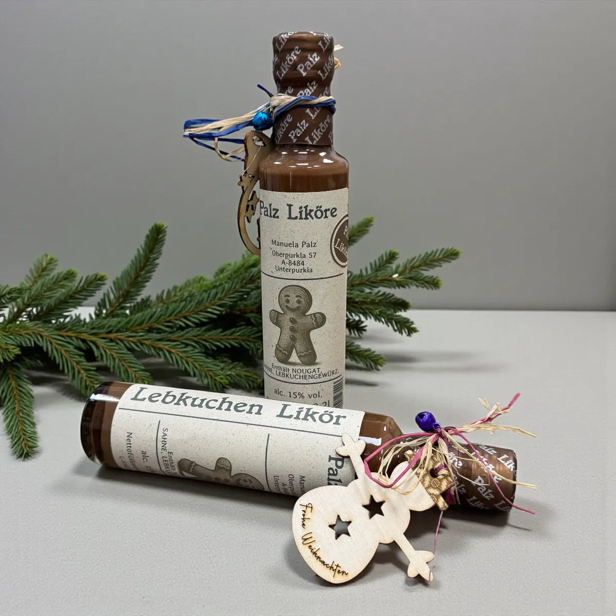 Lebkuchen Likör 0,2 l mit braunem Holzverschluss, Lebkuchen-Holzanhänger und blauem Band, umgeben von Fichtenzweigen und…