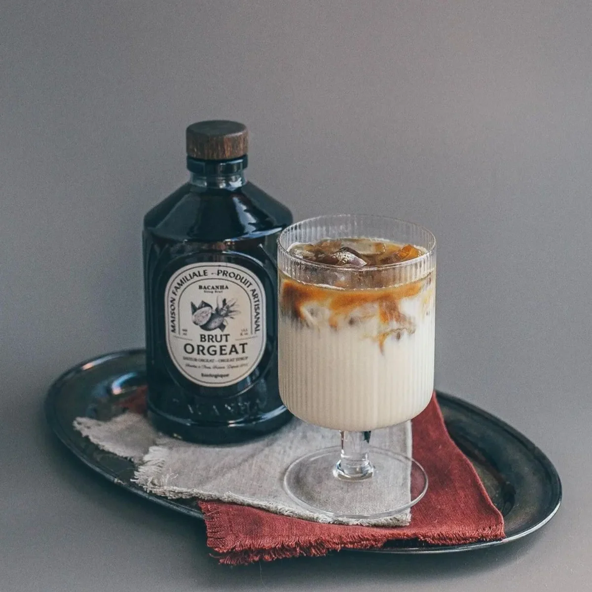Dunkle Flasche Orgeat-Sirup neben einem Cocktail mit Milchschaum und braunem Sirup auf schwarzem Tablett