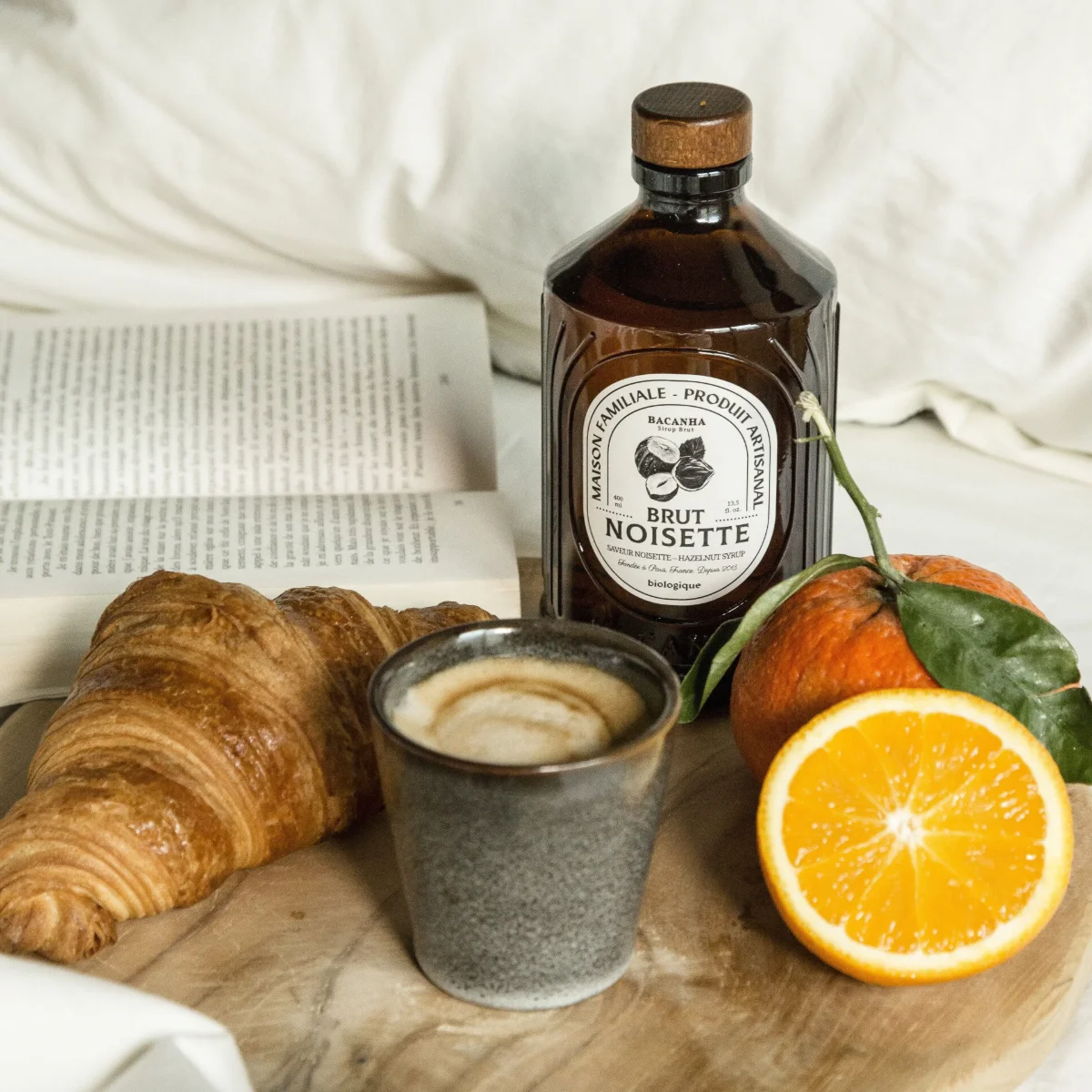 Haselnusssirup in dunkler Flasche mit Korkdeckel neben Cappuccino, Croissant und Orangenscheibe auf Holzbrett