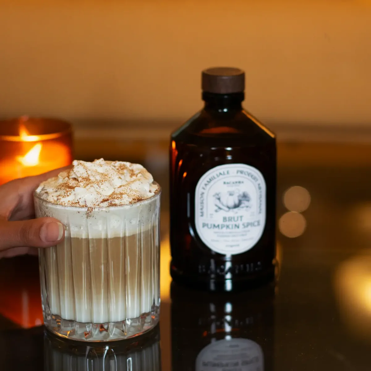 Pumpkin Spice Sirup von BACANHA in dunkler Glasflasche neben einem Glas mit cremigem Getränk, bestreut mit Zimtpulver, im…