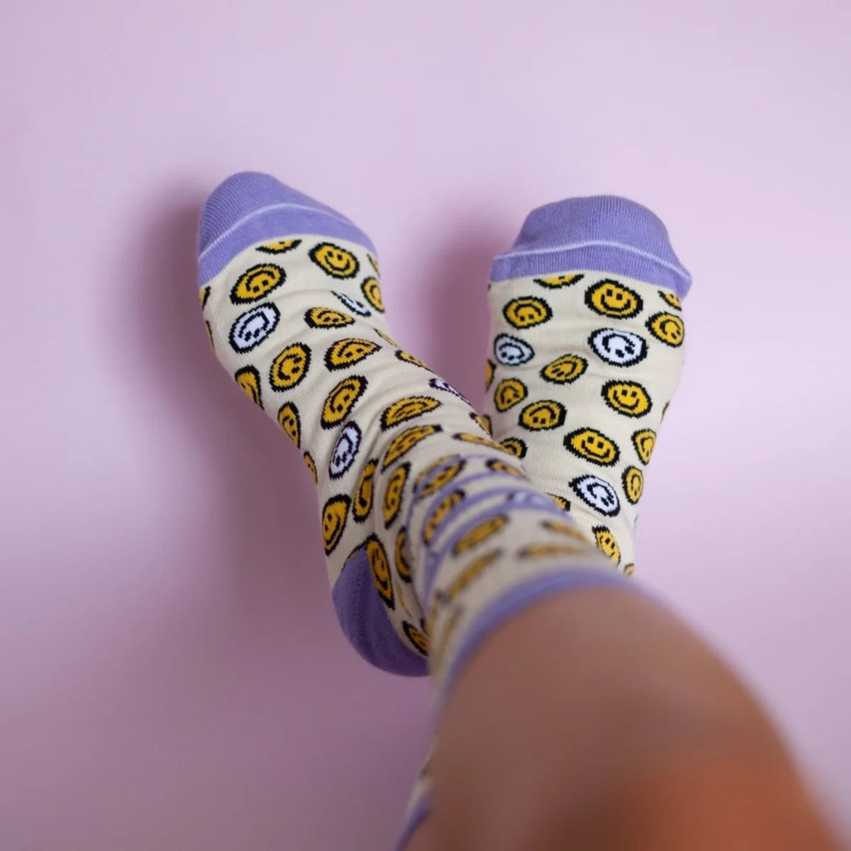 Zwei Sportsocken an Füßen, von oben fotografiert: cremefarbenes Schaftteil mit gelben und blauen Smiley-Motiven, lila…