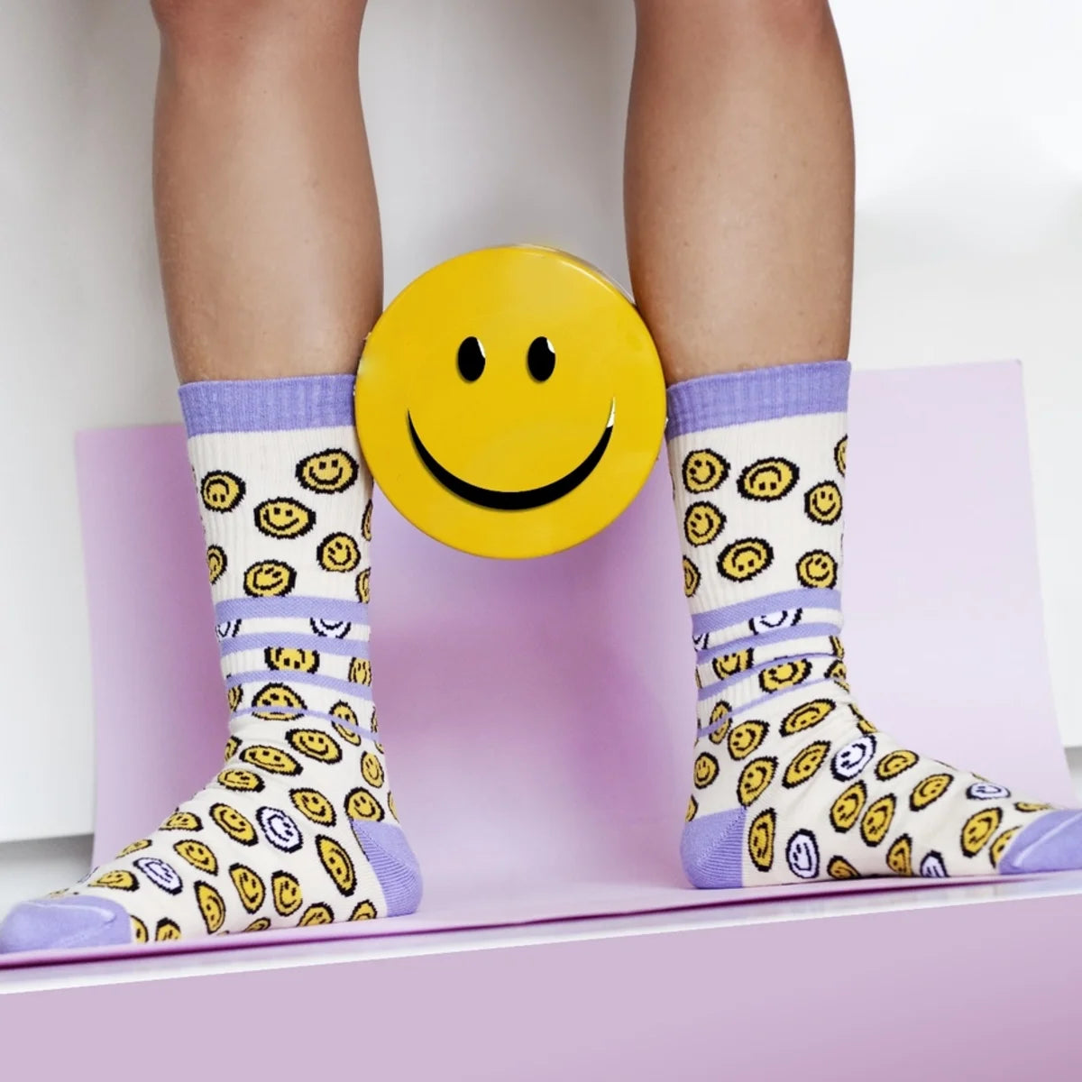 Sportsocken mit gelben Smiley-Motiven auf Creme und lila Streifen, getragen an den Beinen, dahinter großes gelbes…