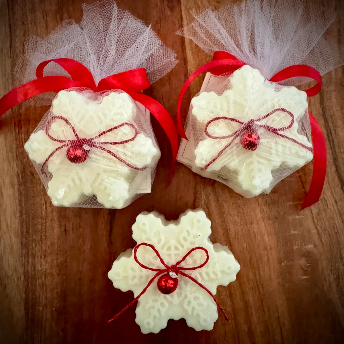 Drei cremeweiße Schneeflocken-Kerzen mit roten Schleifen und Glöckchen, zwei in Tüll verpackt, auf Holzuntergrund.