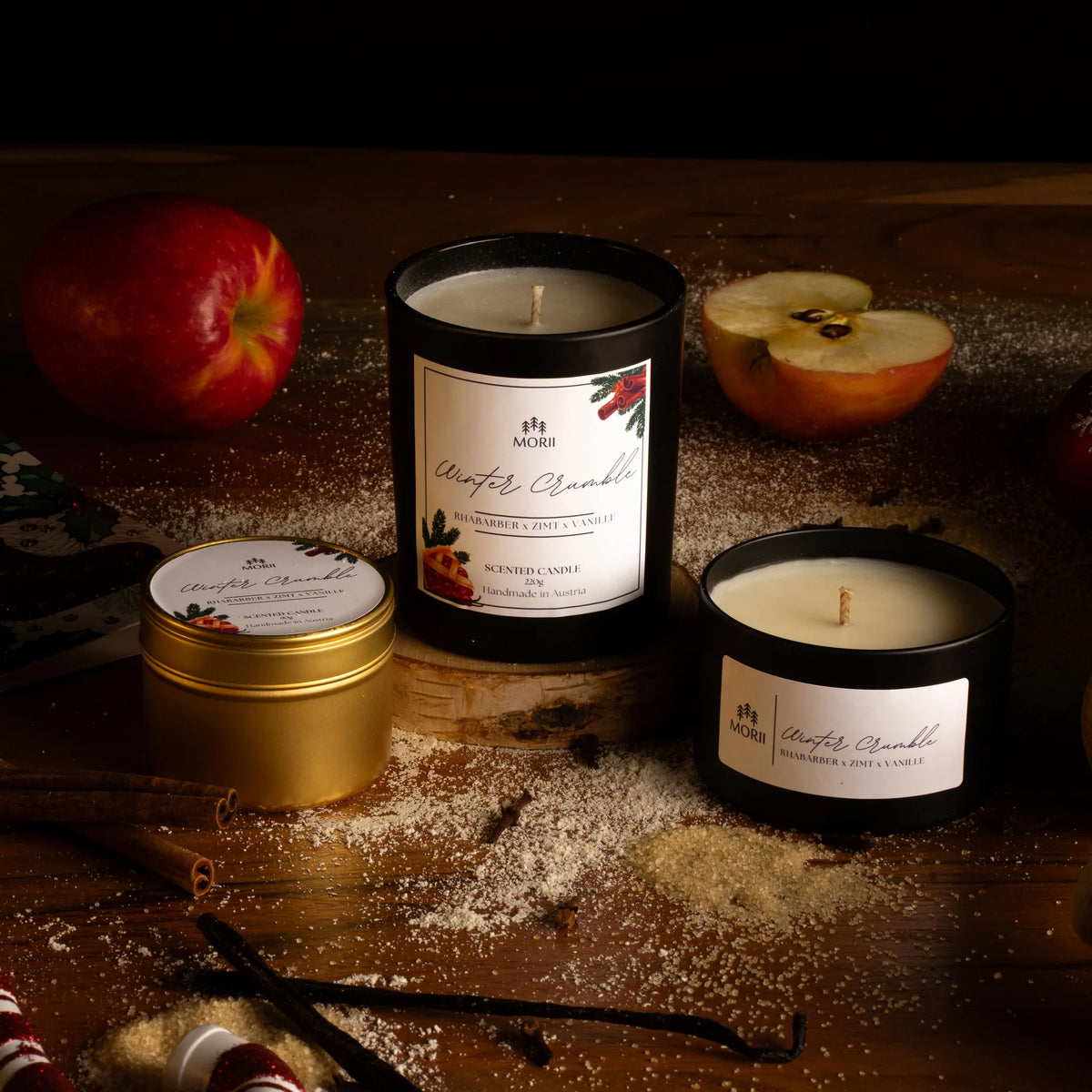 Drei Duftkerzen und eine Dose von Morii arrangiert auf dunklem Holz mit Puderzucker, frischen Äpfeln und Vanilleschoten…