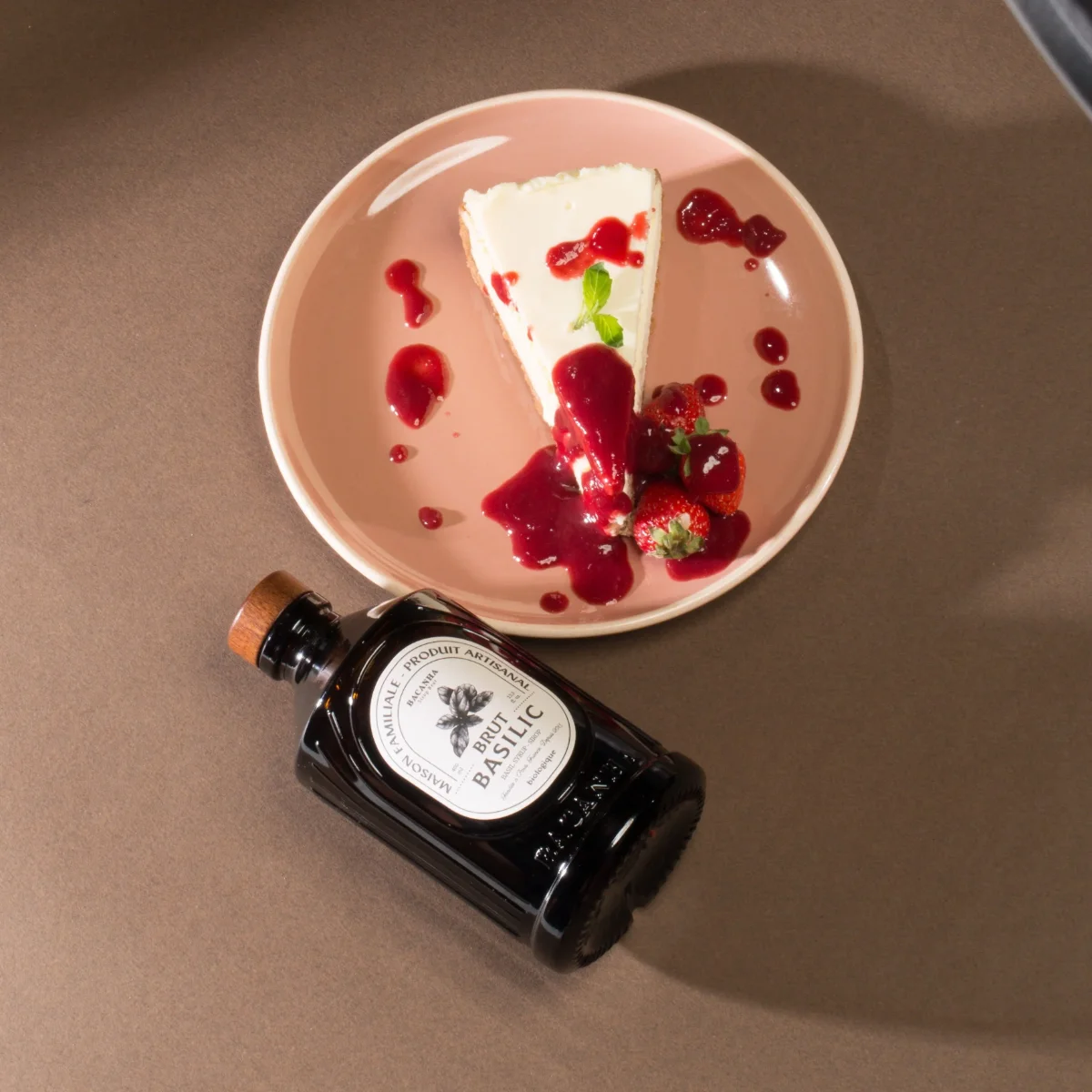 Schwarze Glasflasche mit Korkverschluss neben rosa Teller mit Käsekuchen, roten Beeren und Basilikumblatt