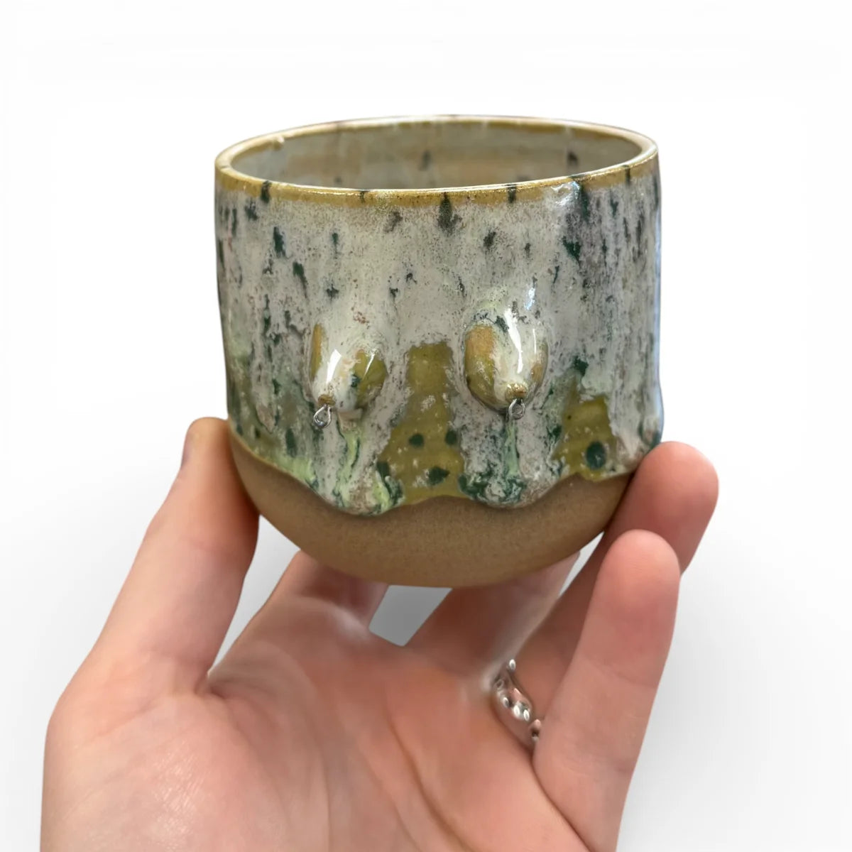 Keramikbecher in der Hand, grün-goldene Glasur mit Sprenkeln, drei modellierte Brüste in Grüntönen, oben heller Rand.