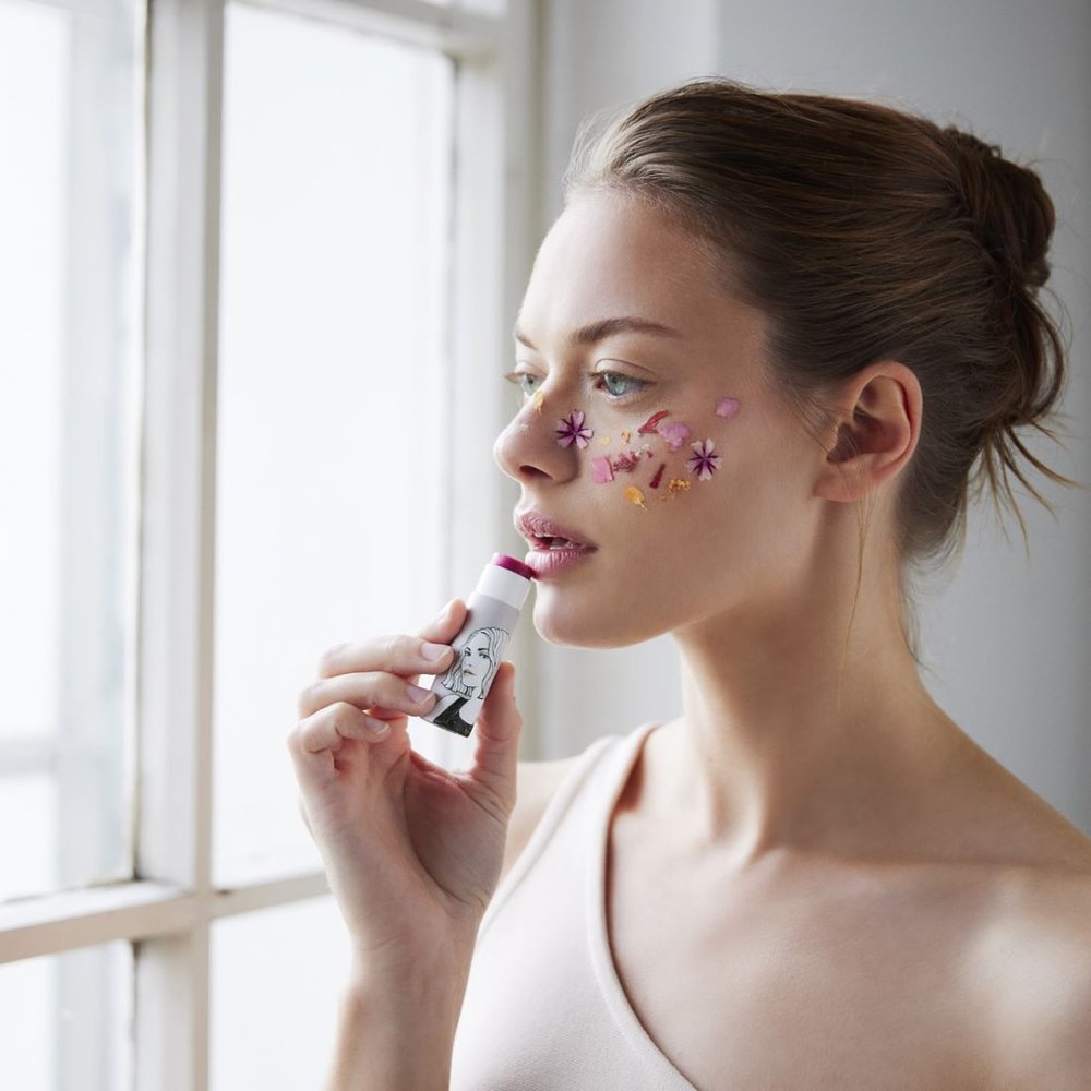 Frau trägt getönten Lipbalm auf, zartes Rosa auf den Lippen, bunte Punkte und Sterne auf der Wange.