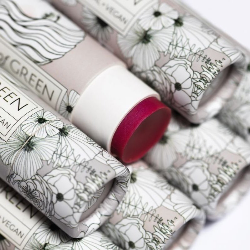 Getönter Lipbalm mit tiefrotem Farbton, umgeben von mehreren Kartonverpackungen mit botanischem Blütenmuster in Schwarz-Weiß.