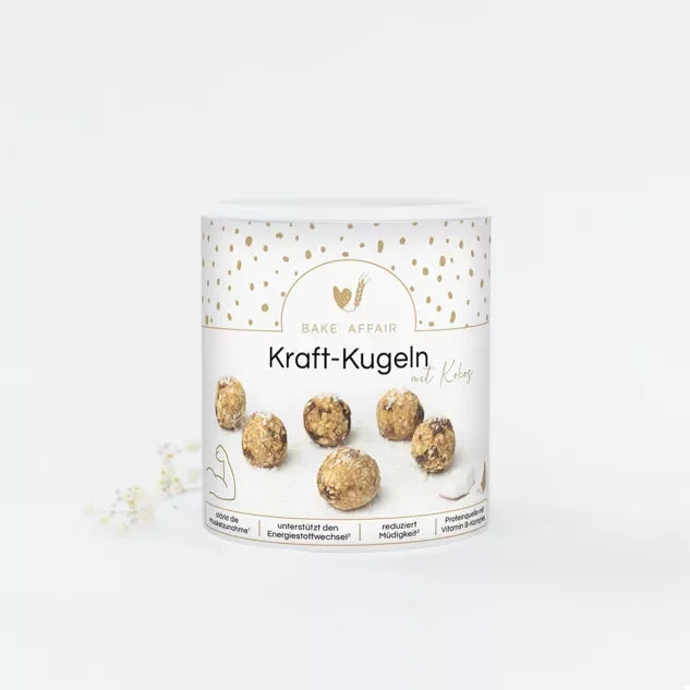 Energiekugeln Kokos: Verpackung von Bake Affair mit sechs goldbraun gesprenkelten Kugeln, daneben weiße Blüten auf hellem…