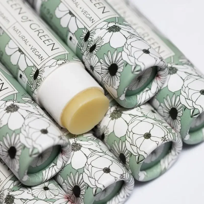 Mehrere grün-weiß bedruckte Lippenbalsam-Hülsen mit Blumenmuster liegen dicht gestapelt; eine ist geöffnet, der gelbliche…