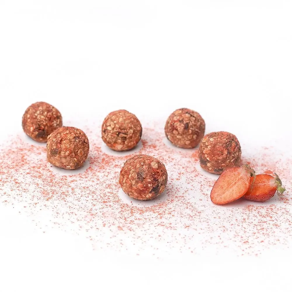 Sechs braune Energiekugeln auf rosa Erdbeergranulat, daneben zwei frische Erdbeeren.