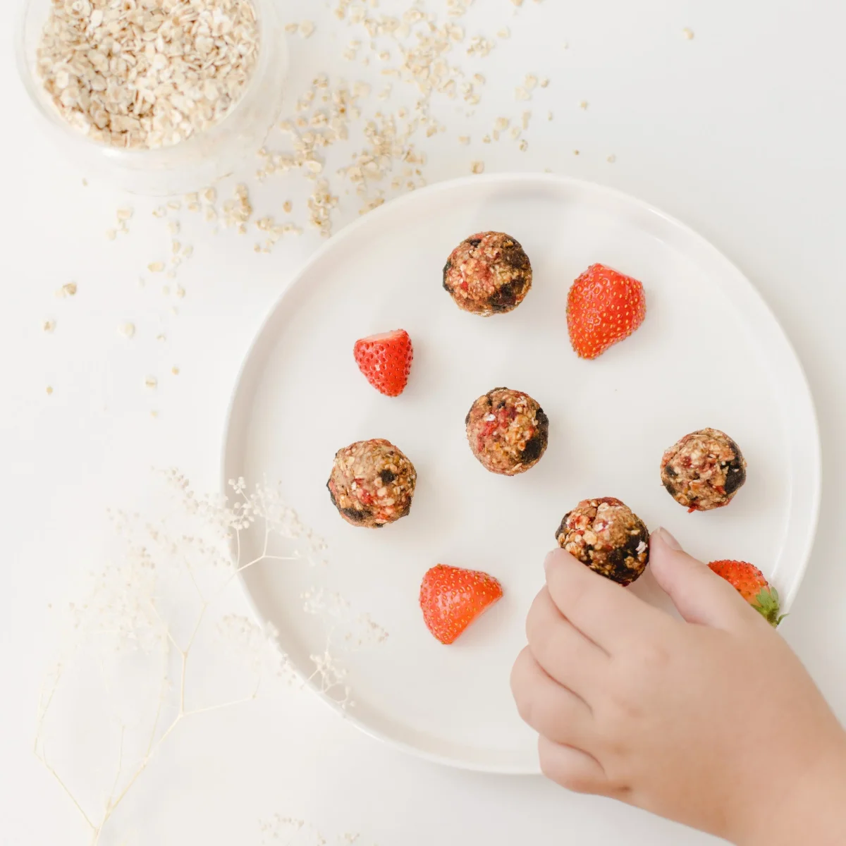 Kinderhand greift nach einer Energiekugel auf weißem Teller mit Erdbeeren und Haferflockenkugeln.