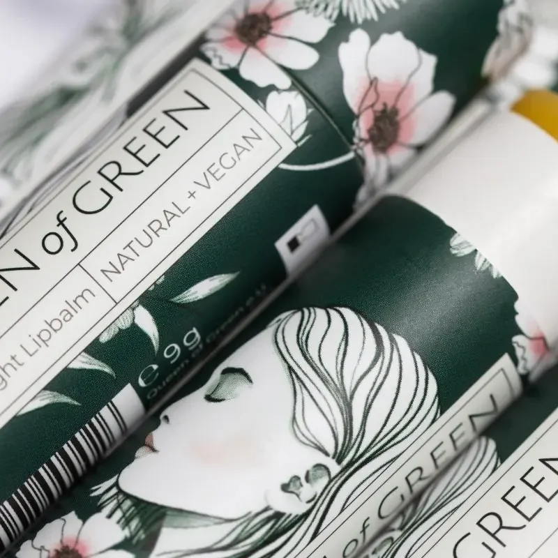 Nahaufnahme zweier dunkelgrüner Lippenpflege-Stifte mit Blumenillustration und einer gezeichneten Frau mit langem Haar…