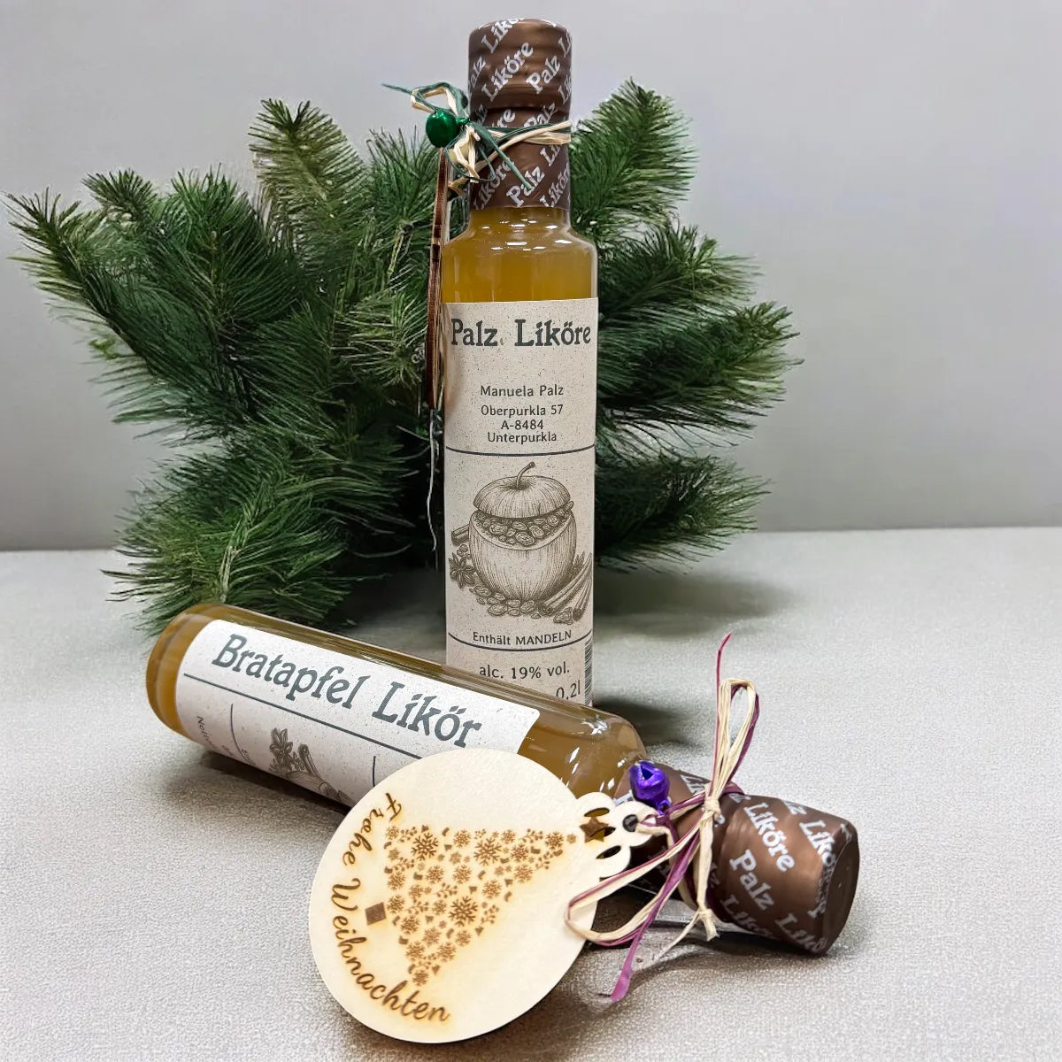 Bratapfel Likör von Palz Liköre: Flasche mit Etikett neben kleiner Flasche, umgeben von Tannenzweigen, Weihnachtskugel und…