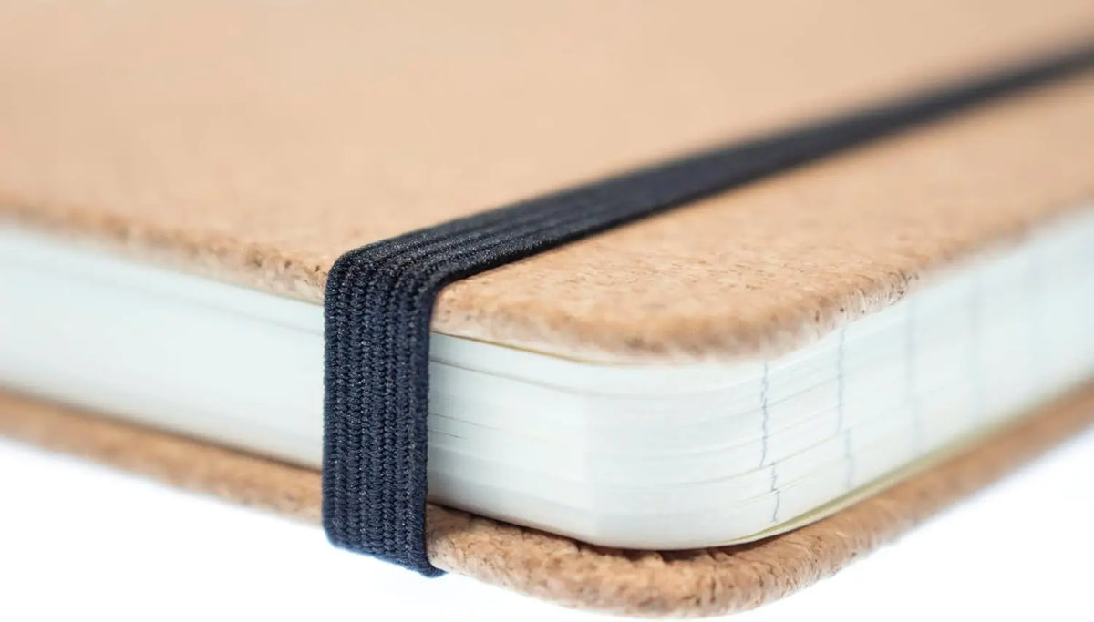 Aufgeschlagenes A5-Notizbuch mit Korkeinband, schwarzes Gummiband-Verschlussband sichtbar, weiße Seiten im Schnitt.
