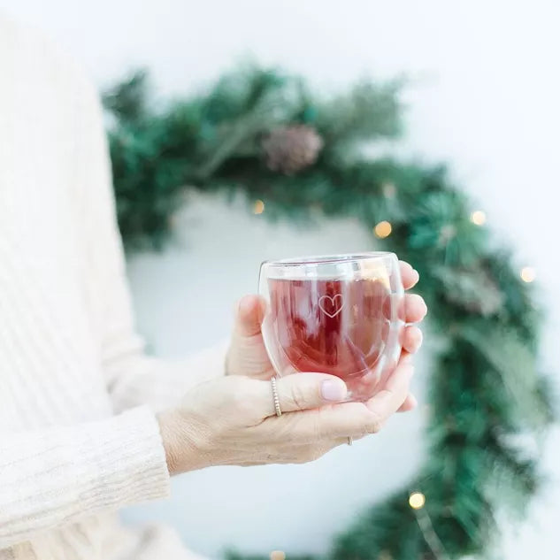 Hände halten ein Glas mit dunkelrotem Tee, im Hintergrund verschwommener Adventskranz mit grünen Zweigen und goldenen…
