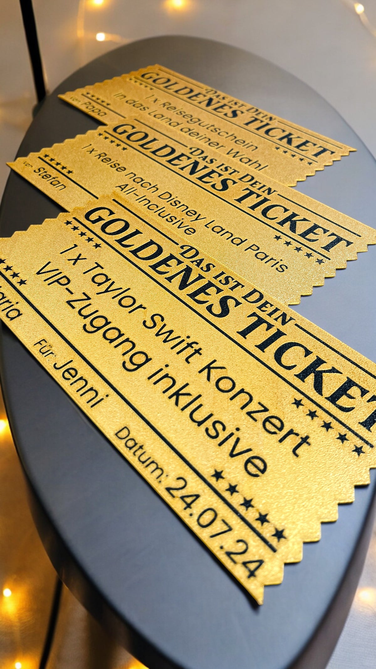 Drei goldene Tickets mit schwarzem Text, gestaffelt auf grauem Untergrund, mit personalisierten Angaben wie Namen, Datum…