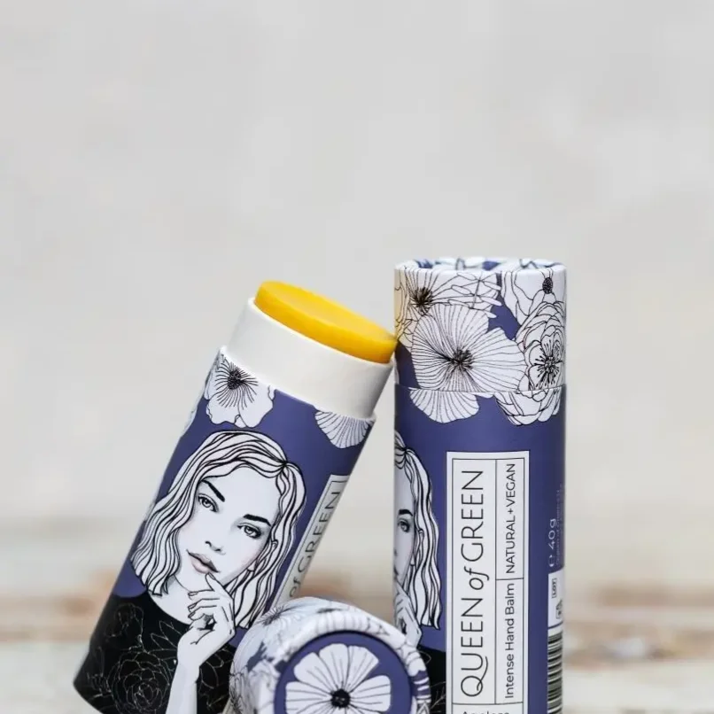 Zwei violette Kartonhülsen mit Blumenzeichnungen und Frauenporträt, eine mit gelbem Handcreme-Stick, im Vordergrund ein…