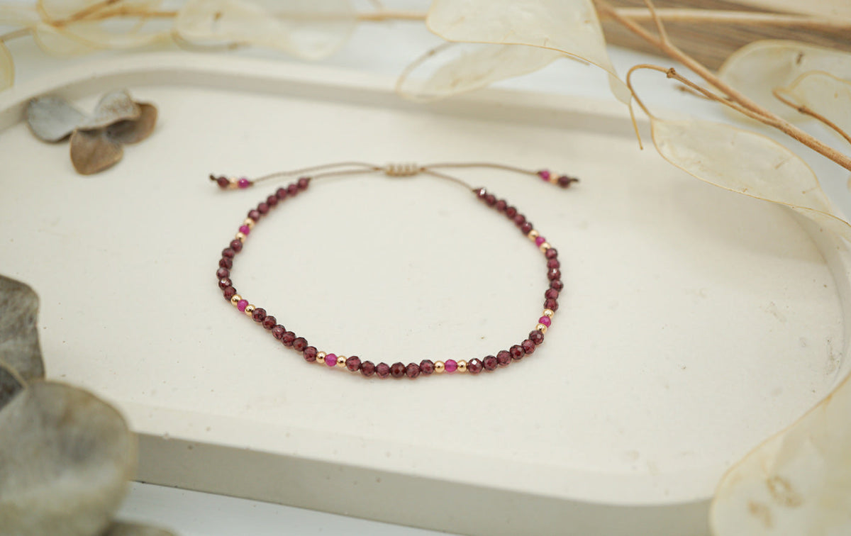 Armband mit roten und rosafarbenen Perlen auf beigem Faden, auf weißer Oberfläche mit getrockneten Blättern.