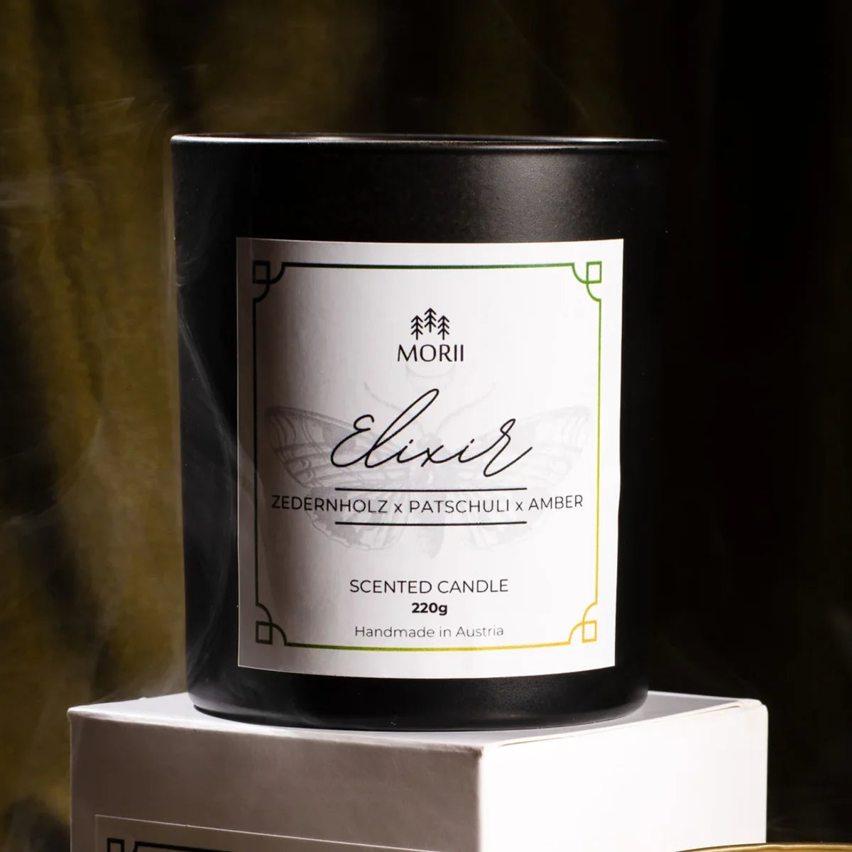 Schwarze Duftkerze mit weißem Label, Aufschrift „Morii Elixir Zedernholz × Patschuli × Amber 220g Handmade in Austria