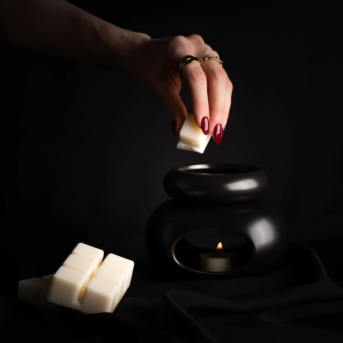 Hand mit rotem Nagellack hält Wachsmelt über schwarze Duftlampe mit brennender Kerze, zwei weitere Melts daneben.