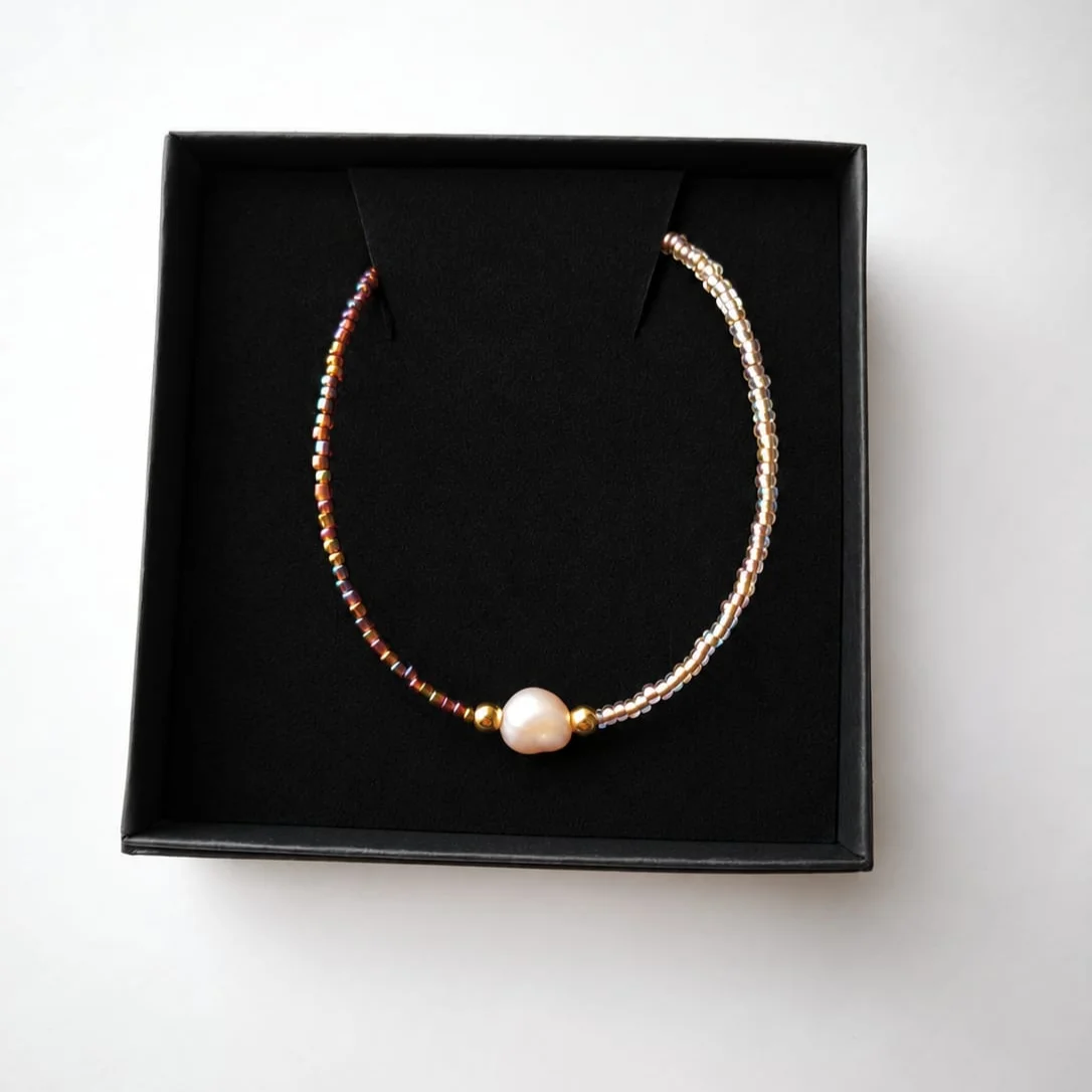 Perlenkette aus kleinen bunten und goldenen Glasperlen mit einer hellen, unregelmäßigen Süßwasserperle in der Mitte, auf…