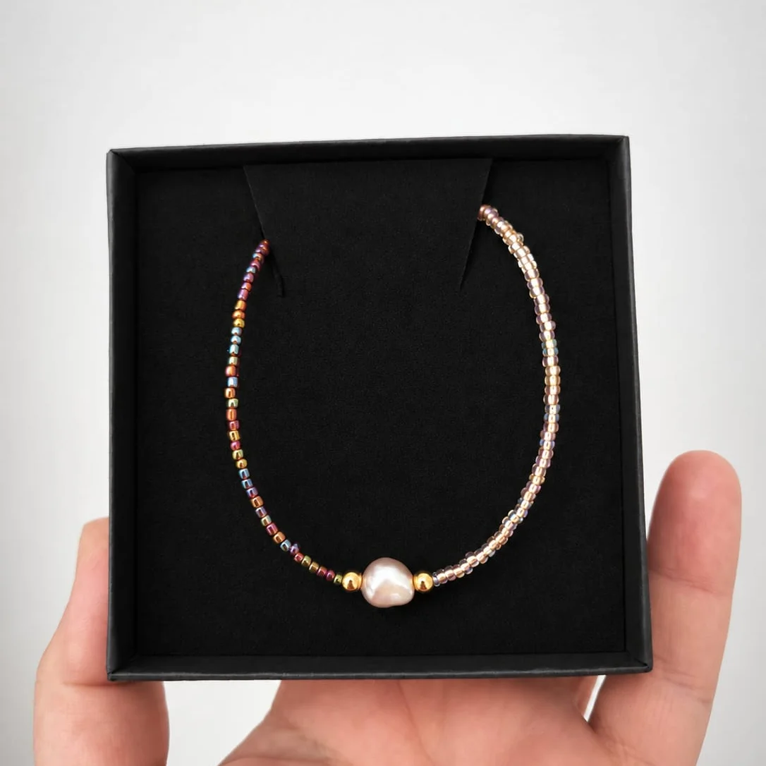 Perlenkette aus kleinen bunten Glasperlen in Rot und Gold, mit einer einzelnen rosafarbenen Süßwasserperle in der Mitte…