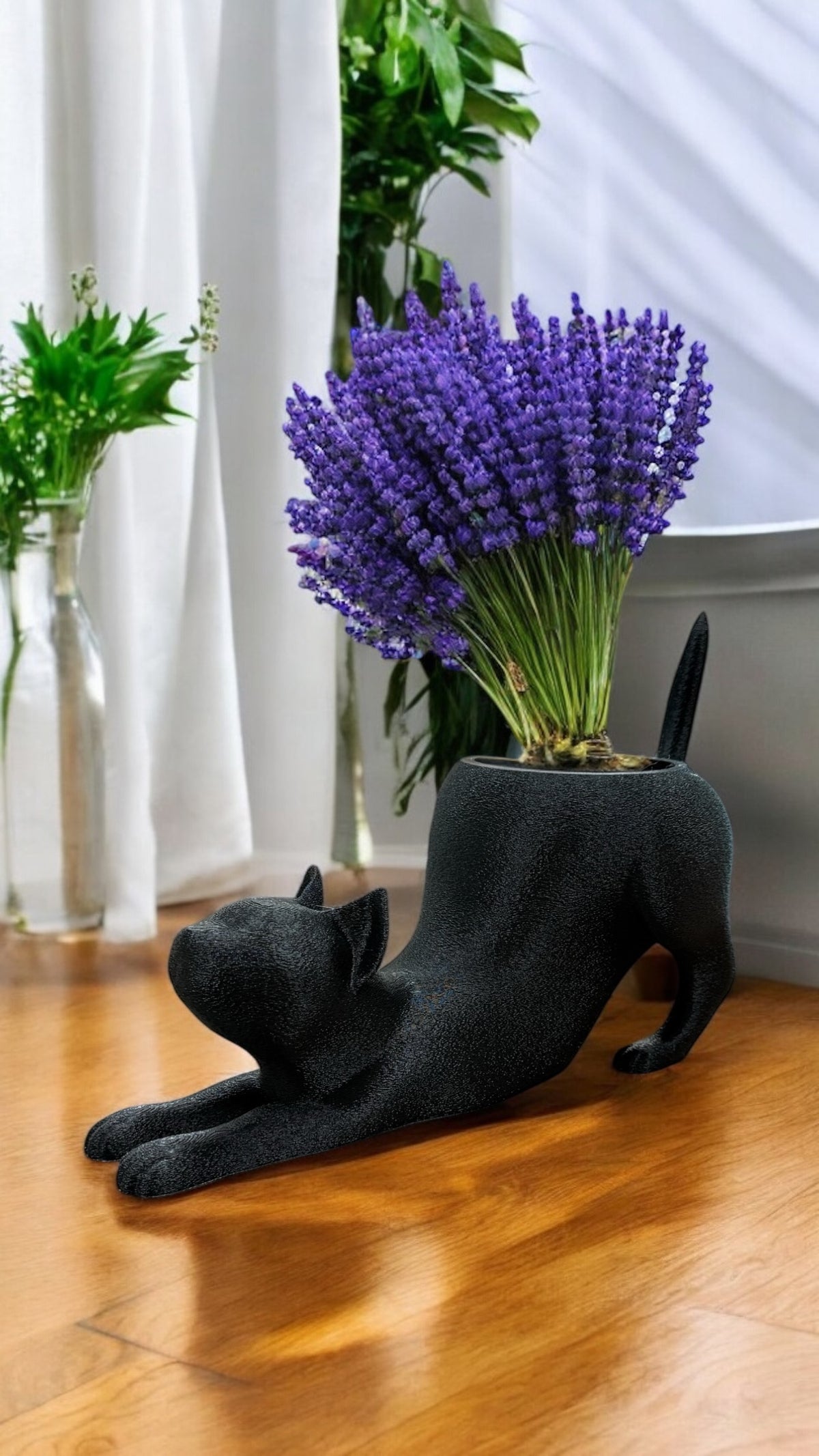 Schwarzer Katzentopf in Streckposition auf Holztisch, gefüllt mit violettem Lavendel, daneben Glasvase mit Grünpflanzen.