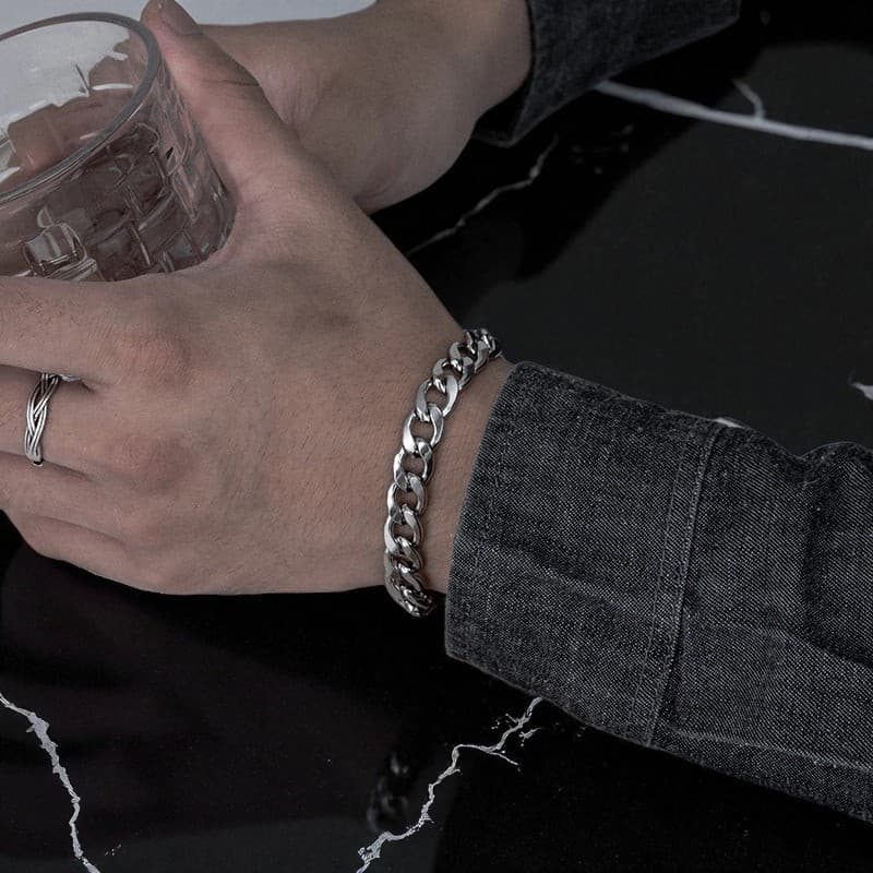 Silbernes Gliederkettchen-Armband am Handgelenk getragen, zentrale Plakette sichtbar, unter grauem Stoff.