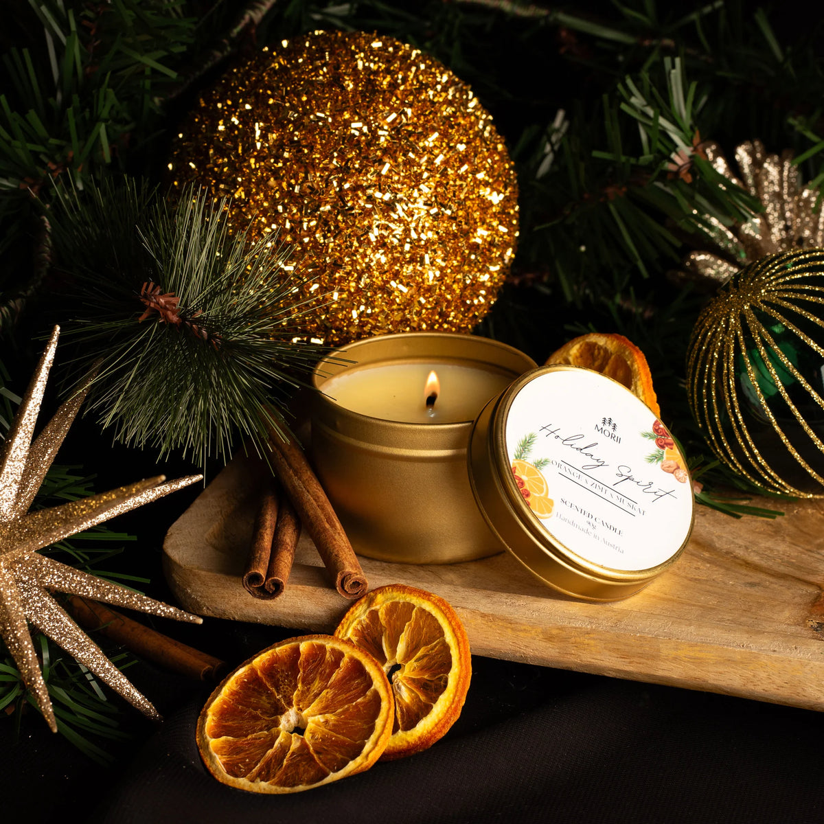 Goldene Duftkerze in Metalldose mit brennender Flamme, daneben getrocknete Orangenscheiben und Zimtstangen auf…