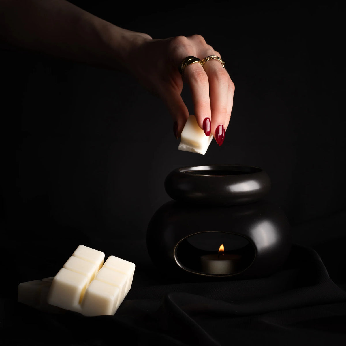 Hand mit roten Nägeln hält Wachsmelt über schwarze Duftlampe mit brennender Flamme, zwei weitere Melts daneben.