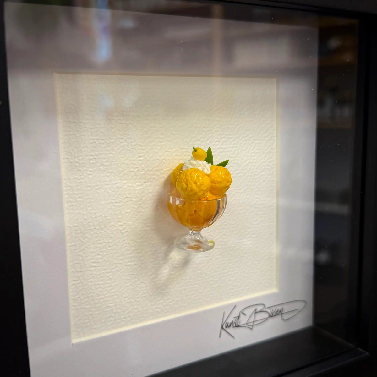 Miniatur-Sorbet aus orangefarbenen Kugeln in Glasschale mit grünem Blatt, auf cremefarbenem Passepartout im schwarzen…