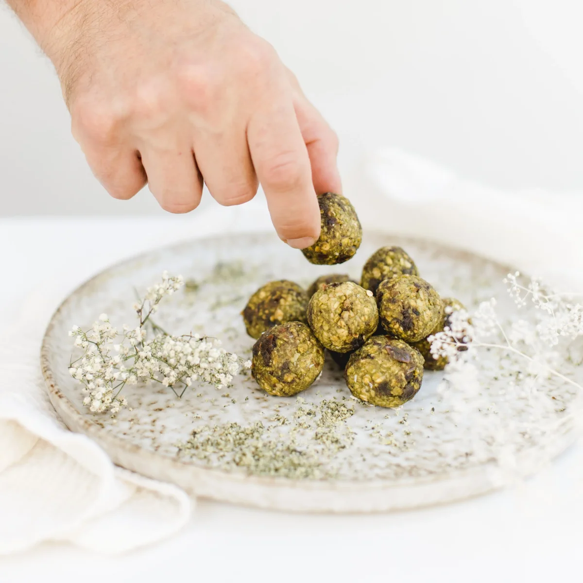 Hand nimmt grüne Energiekugel von Keramikteller mit Matcha-Pulver und Trockenblüten