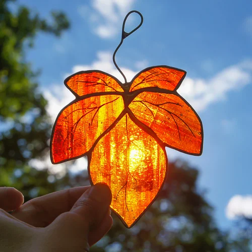 Fensterbild Glas Herbst: Orangerotes Blatt in Tiffany-Technik gegen Himmel, in Hand gehalten, Licht scheint durch.