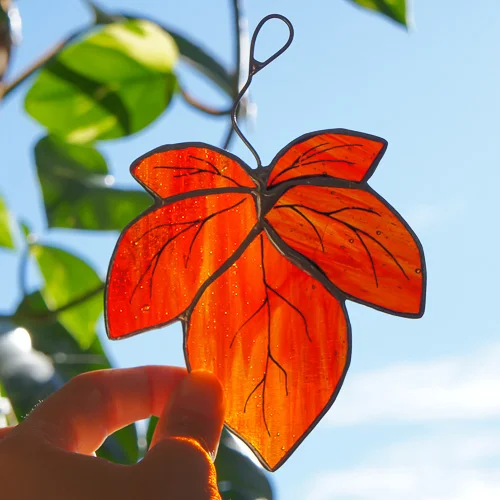 Orangerotes Glasblatt mit handgemalten Wurzeln, gegenlicht vor blauem Himmel und grünem Laub, an Metallöse hängend.