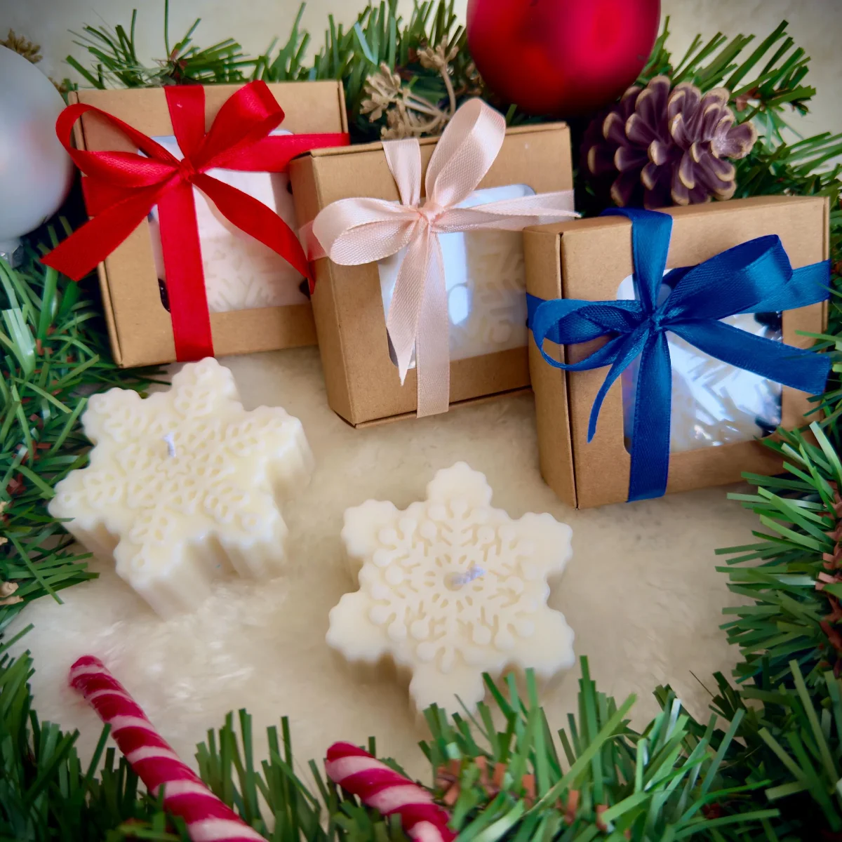 Schneeflocken-Kerze aus Sojawachs in Creme neben drei Kraftpapierboxen mit roten, beigen und blauen Schleifen, umgeben von…