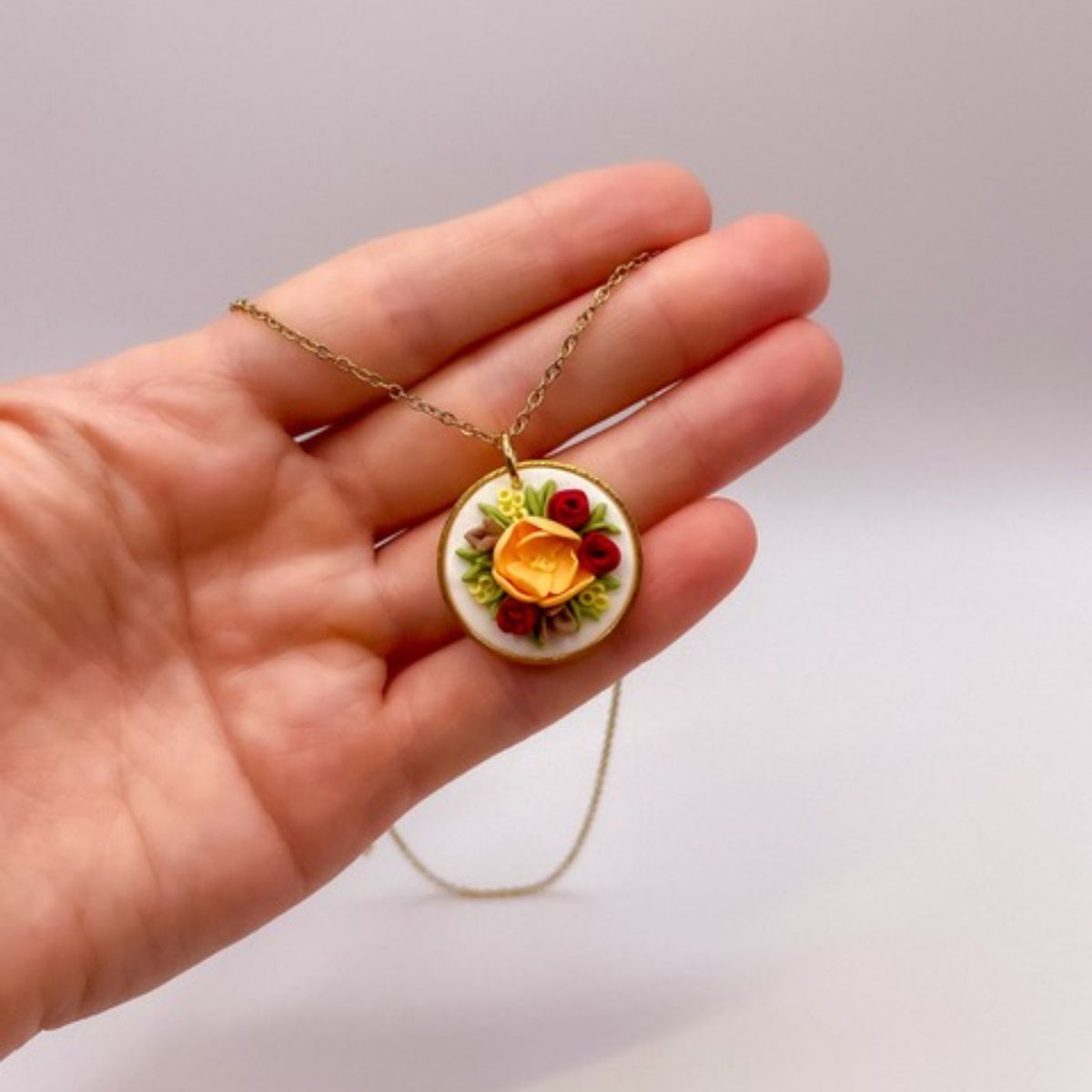 Halskette in der Hand: Goldkette mit rundem Anhänger, darauf gelbe und rote Blüten auf cremefarbenem Grund.