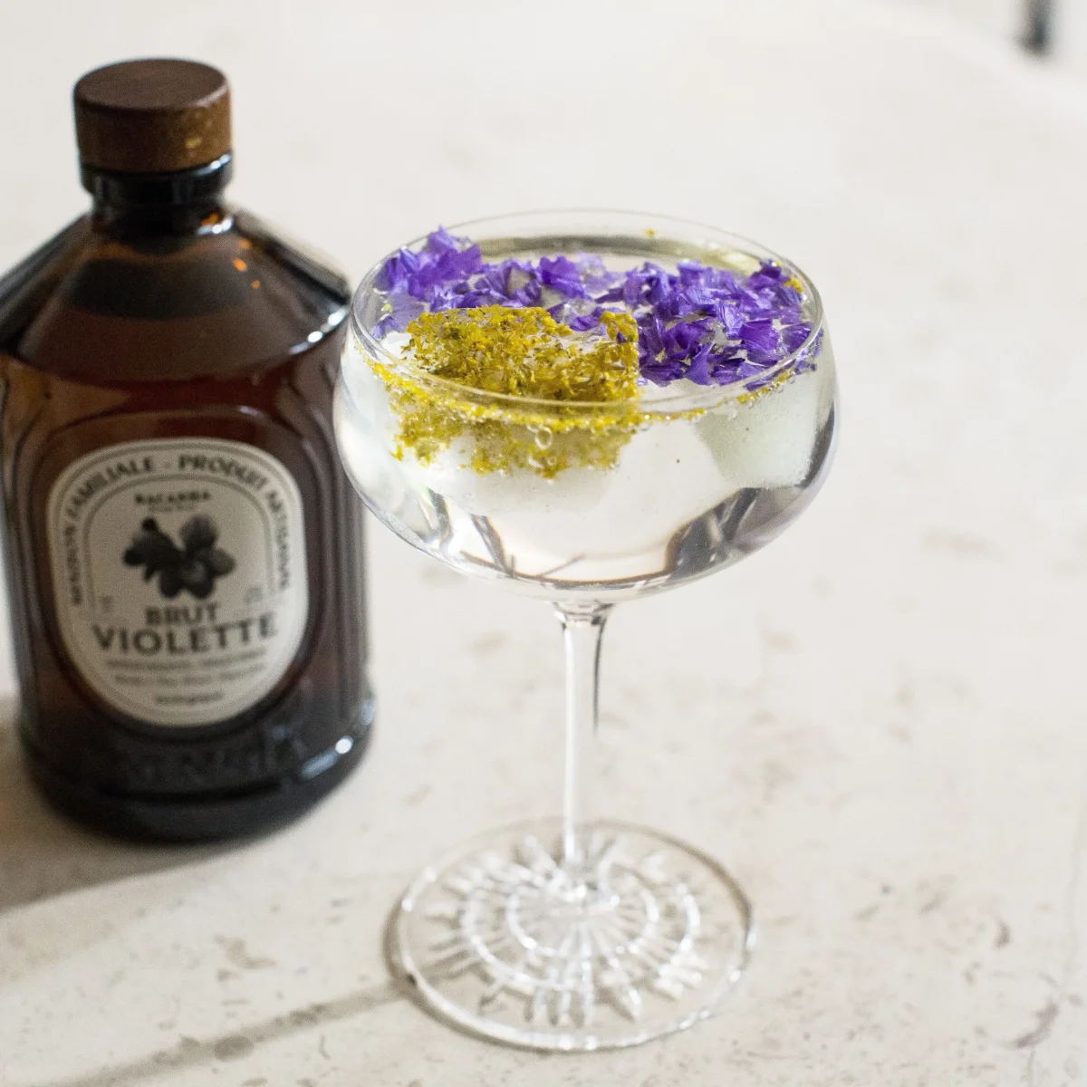 Veilchensirup in brauner Flasche neben einem Cocktail mit violetten Blüten und gelbem Zucker am Glasrand