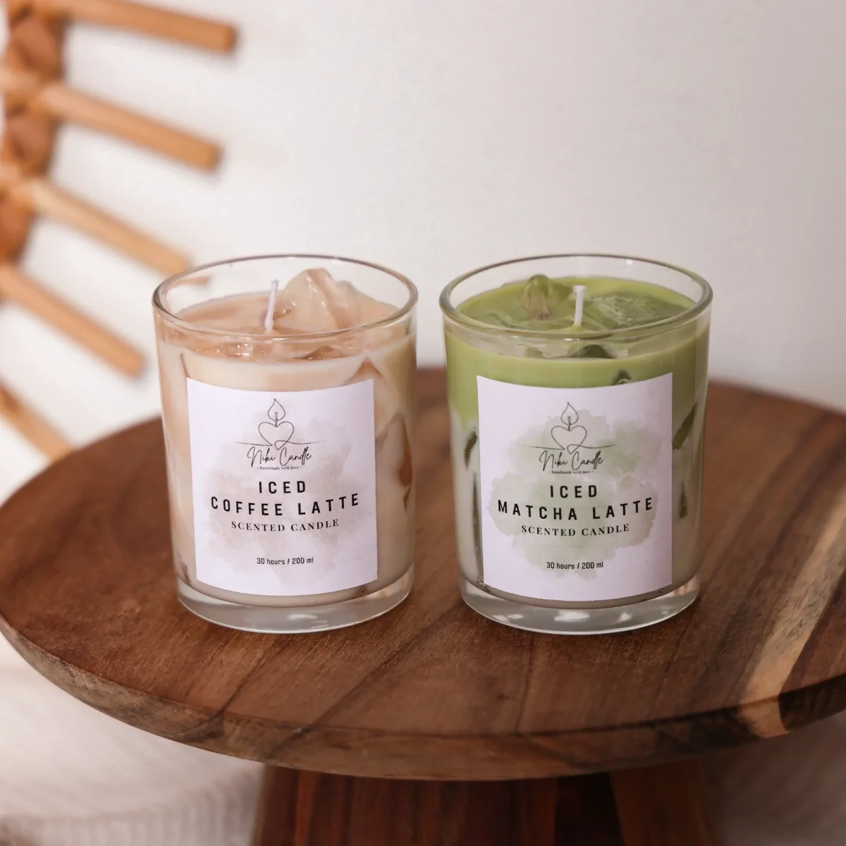 Zwei Duftkerzen nebeneinander auf rundem Holztisch: links Iced Coffee Latte in Beige mit Eiswürfeln, rechts Iced Matcha…