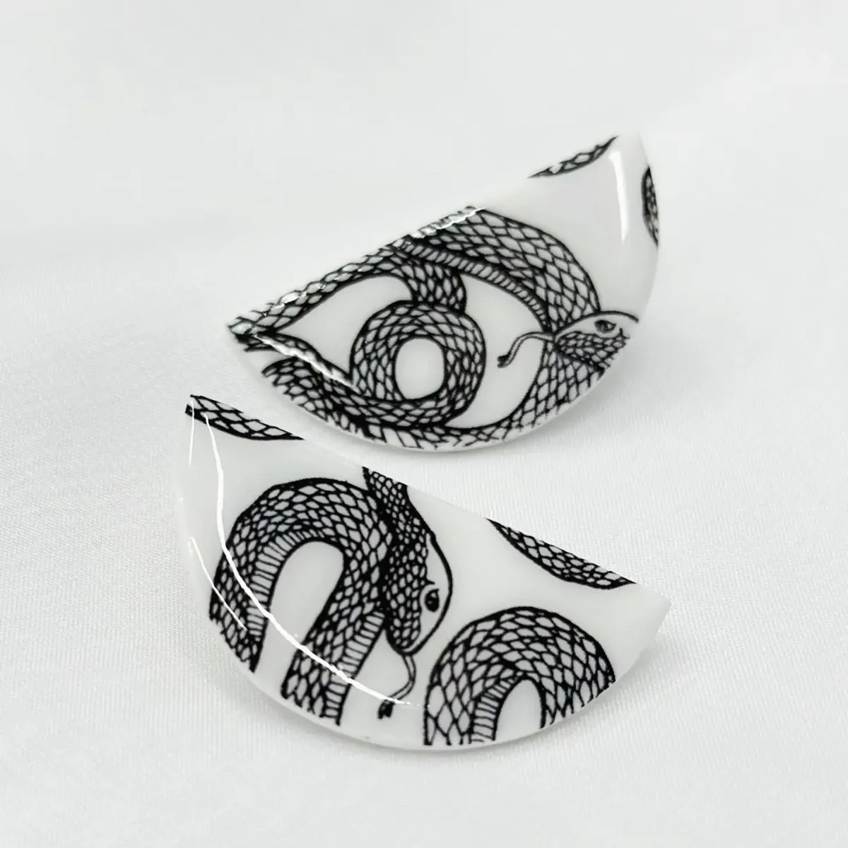 Zwei flache Ohrstecker aus Kunststoff mit schwarzen Schlangen-Motiven auf weißem Grund, Draufsicht