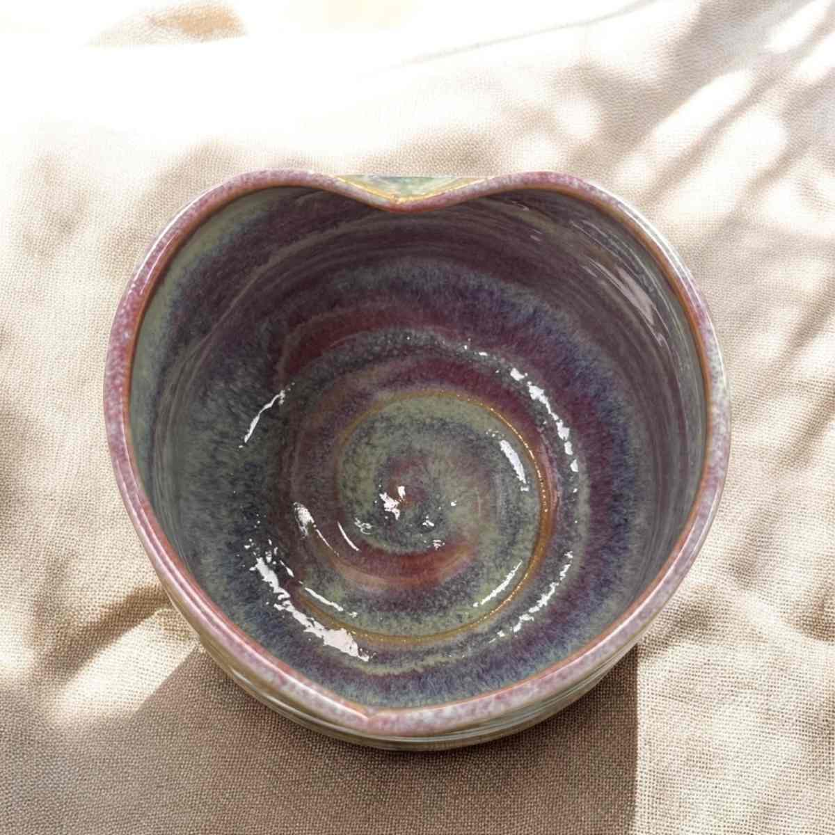Herzförmige Tasse von oben, Innenansicht mit lila-grauer Glasur in Farbverläufen, heller Stoff im Hintergrund.
