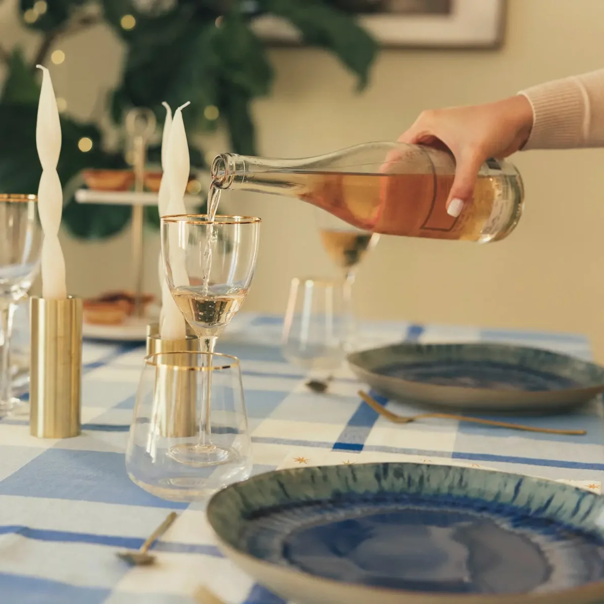 Gedeckter Tisch mit blau-weiß kariertem Tuch: Hand gießt Rosé in ein Glas, daneben blau glasierte Teller mit Goldbesteck…