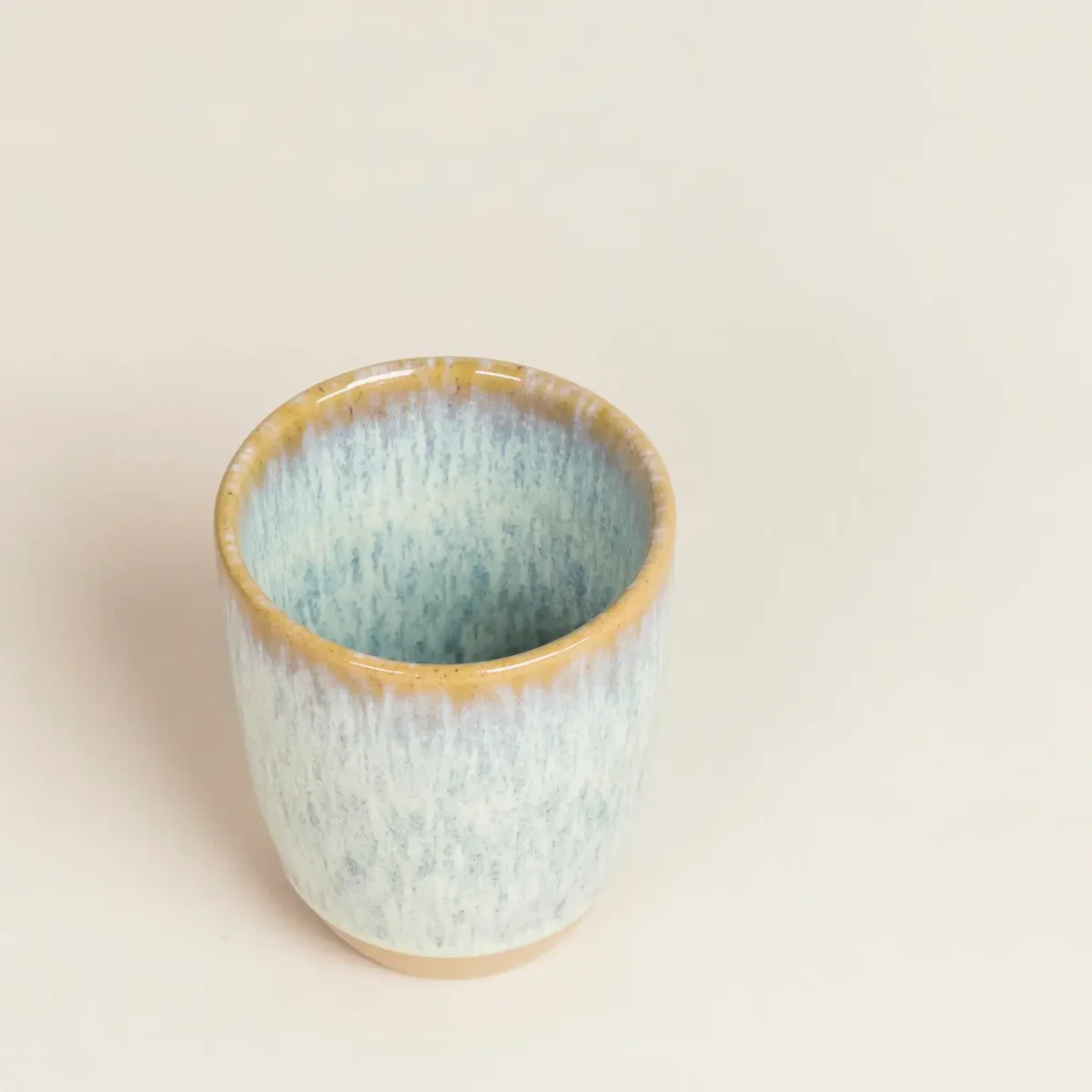 Blick von oben in eine becherförmige Keramiktasse mit hellblau-grüner, leicht gesprenkelter Glasur und gelblichem Rand, auf…