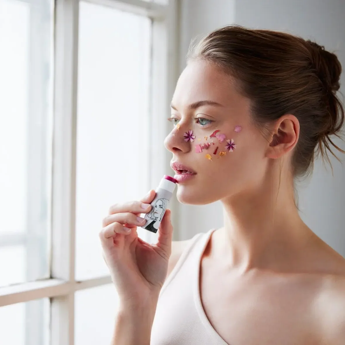 Frau mit zusammengebundenem Haar vor einem Fenster trägt mit einem Lippenstift-Stift Farbe auf die Lippen auf; auf ihrer…